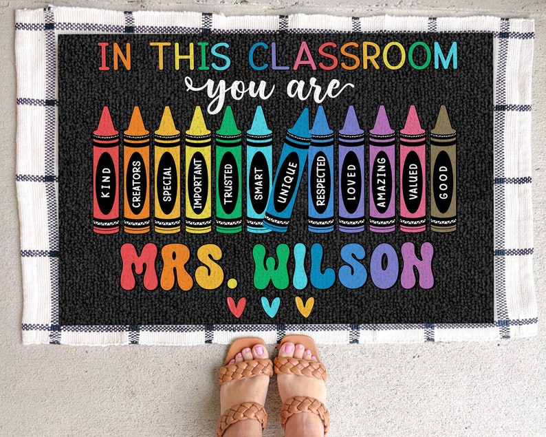 May include: A black and white welcome mat with the text "In this classroom you are" and a row of colorful crayons with words like "kind", "creators", "special", "important", "trusted", "smart", "unique", "respected", "loved", "amazing", "valued", and "good" written on them. The mat also has the text "Mrs. Wilson" in colorful letters.