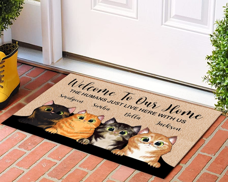 May include: Welcome To Our Home doormat with a brown background and the text "Welcome To Our Home The Humans Just Live Here With Us" with the names Smidgeon, Simba, Bella, and Jackson. The doormat features four cartoon cats with different colored fur.
