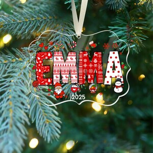 May include: A clear, rectangular Christmas ornament with the name "EMMA" in red and white festive patterns. The year "2025" is printed below the name, along with holiday-themed illustrations like Santa, a snowman, and candy canes.