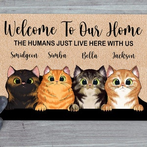 May include: Welcome To Our Home doormat with a brown burlap background. The text "THE HUMANS JUST LIVE HERE WITH US" is printed in white. Four cats are peeking over the top of the doormat. The cats are named Smidgeon, Simba, Bella, and Jackson.
