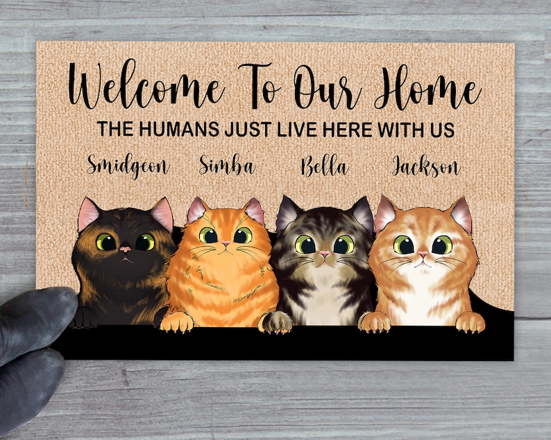 May include: Welcome To Our Home doormat with a brown burlap background. The text "THE HUMANS JUST LIVE HERE WITH US" is printed in white. Four cats are peeking over the top of the doormat. The cats are named Smidgeon, Simba, Bella, and Jackson.