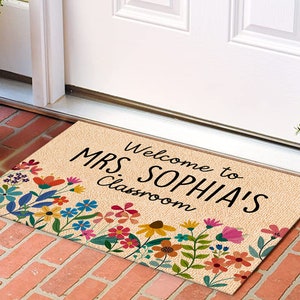 Może przedstawiać: Prostokątna wycieraczka z beżowym tłem i wielokolorową kwiatową obw&oacute;dką. Tekst "Welcome to MRS. SOPHIA'S Classroom" jest wydrukowany na czarno. Mata jest umieszczona na ceglanej powierzchni.