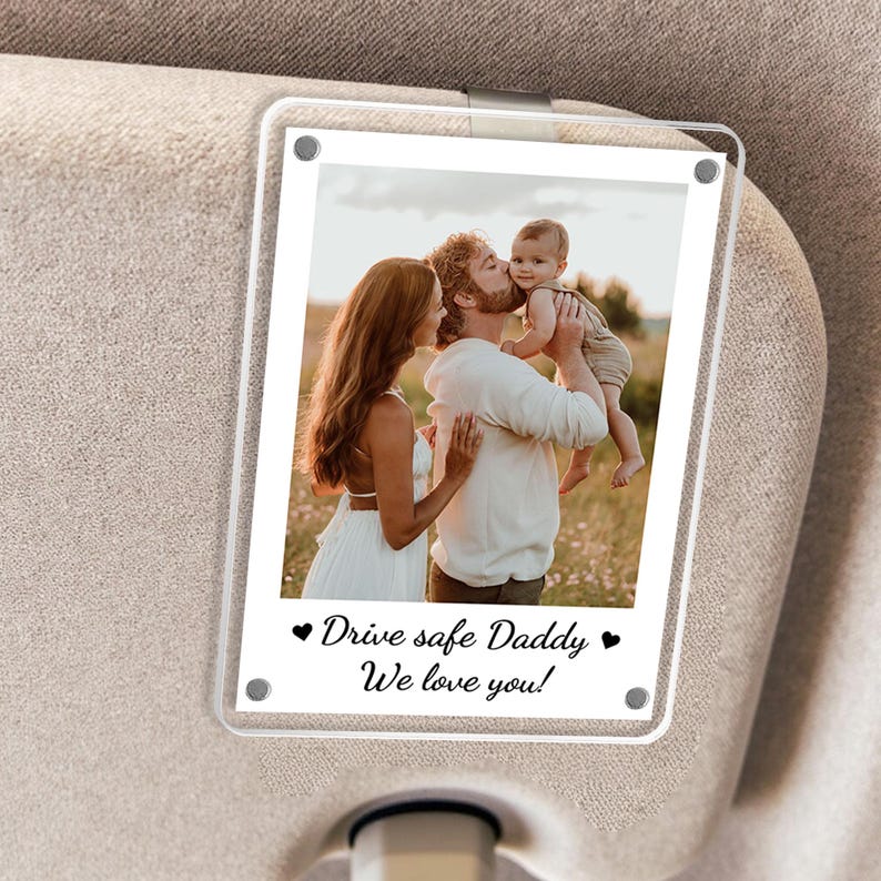 May include: A clear acrylic car visor photo holder with a family photo. The photo shows a family of three in a field. The text on the holder reads "Drive safe Daddy, We love you!" in black script. The holder is attached to a car visor.