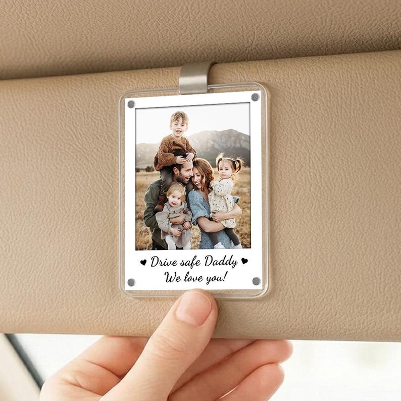 May include: A clear, rectangular car visor photo holder with a family photo. The photo features a family of five, with the text "Drive safe Daddy We love you!" in black. The holder is attached to a beige car visor.