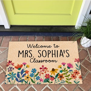 Może przedstawiać: Prostokątna wycieraczka z napisem "Welcome to MRS. SOPHIA'S Classroom" w kolorze czarnym. Mata ma beżowe tło i jest ozdobiona kolorowymi wzorami kwiatowymi w kolorach czerwonym, pomarańczowym, ż&oacute;łtym, r&oacute;żowym i niebieskim.
