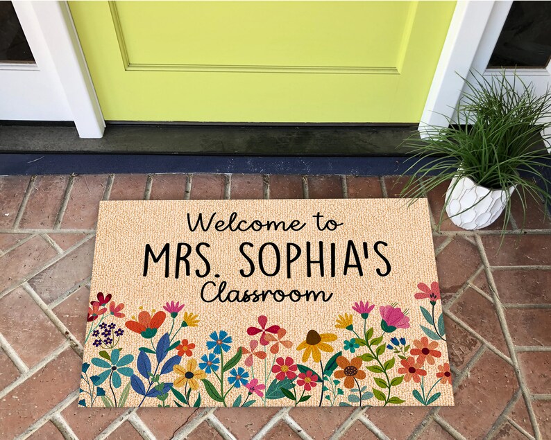 Może przedstawiać: Prostokątna wycieraczka z napisem "Welcome to MRS. SOPHIA'S Classroom" w kolorze czarnym. Mata ma beżowe tło i jest ozdobiona kolorowymi wzorami kwiatowymi w kolorach czerwonym, pomarańczowym, ż&oacute;łtym, r&oacute;żowym i niebieskim.