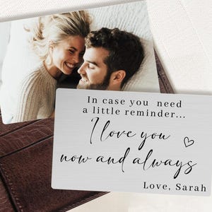 May include: A silver metal wallet card with the text "In case you need a little reminder... I love you now and always Love, Sarah." A photo of a smiling couple is above the card. The card is placed on a brown leather wallet.