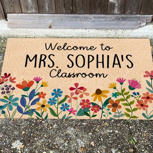 Może przedstawiać: Prostokątna wycieraczka z czarnym napisem "Welcome to MRS. SOPHIA'S Classroom". Mata ma beżowe tło i jest ozdobiona kolorową kwiatową bordiurą z kwiatami w odcieniach niebieskiego, pomarańczowego, r&oacute;żowego i ż&oacute;łtego.