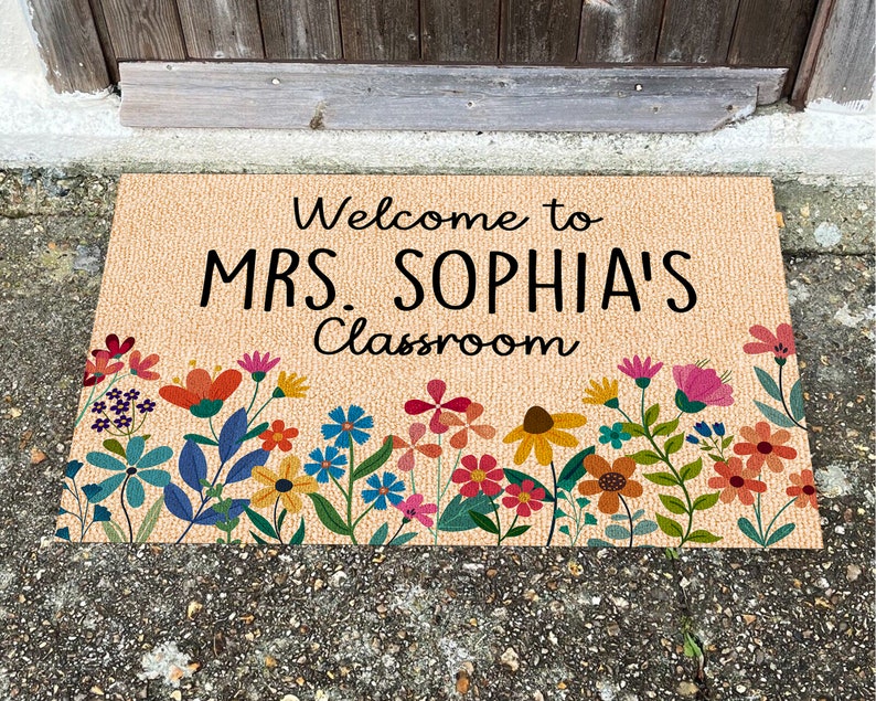 Może przedstawiać: Prostokątna wycieraczka z czarnym napisem "Welcome to MRS. SOPHIA'S Classroom". Mata ma beżowe tło i jest ozdobiona kolorową kwiatową bordiurą z kwiatami w odcieniach niebieskiego, pomarańczowego, r&oacute;żowego i ż&oacute;łtego.