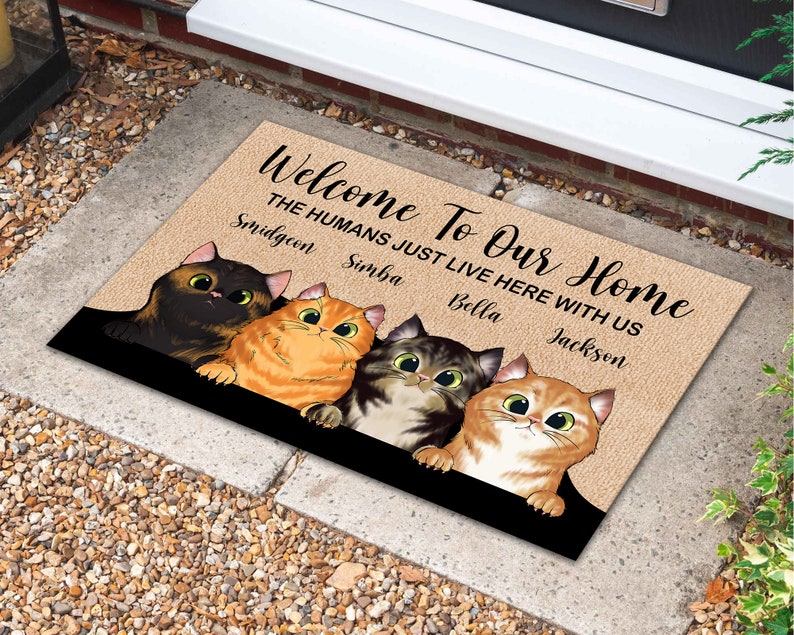 May include: Welcome To Our Home doormat with a brown background and four cats with different colored fur. The text on the doormat reads "Welcome To Our Home The Humans Just Live Here With Us Smidgeon Simba Bella Jackson".