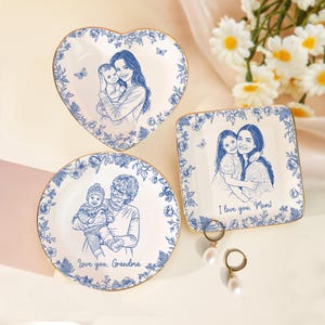 Puede incluir: Tres platos decorativos de cer&aacute;mica con bordes florales azules y bordes dorados. Cada plato presenta un boceto azul de un miembro de la familia sosteniendo a un ni&ntilde;o. Un plato tiene forma de coraz&oacute;n, otro es cuadrado y otro redondo. El texto en los platos dice "I love you, Mom!" y "Love you, Grandma."