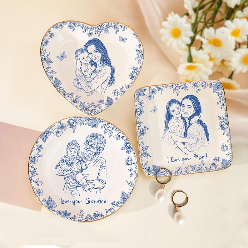 Puede incluir: Tres platos decorativos de cer&aacute;mica con bordes florales azules y bordes dorados. Cada plato presenta un boceto azul de un miembro de la familia sosteniendo a un ni&ntilde;o. Un plato tiene forma de coraz&oacute;n, otro es cuadrado y otro redondo. El texto en los platos dice "I love you, Mom!" y "Love you, Grandma."