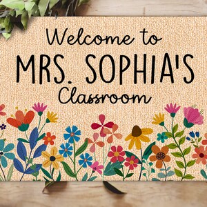 Może przedstawiać: Beżowa wycieraczka z czarnym napisem "Welcome to MRS. SOPHIA'S Classroom". Dolna krawędź zdobiona kolorową kwiatową bordiurą z r&oacute;żnymi kwiatami w odcieniach czerwieni, pomarańczy, r&oacute;żu, błękitu i ż&oacute;łci.