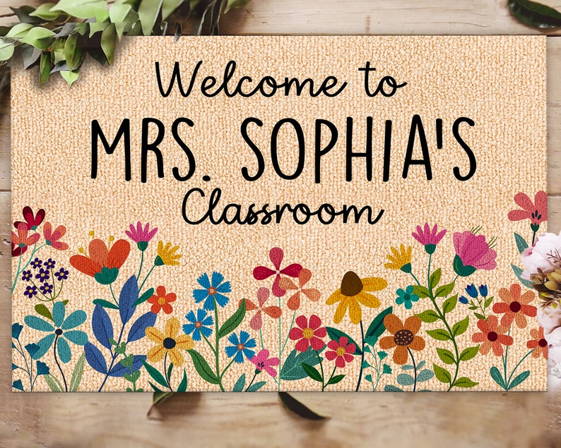 Może przedstawiać: Beżowa wycieraczka z czarnym napisem "Welcome to MRS. SOPHIA'S Classroom". Dolna krawędź zdobiona kolorową kwiatową bordiurą z r&oacute;żnymi kwiatami w odcieniach czerwieni, pomarańczy, r&oacute;żu, błękitu i ż&oacute;łci.