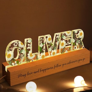 May include: A wooden name sign with the name "OLIVER" spelled out in white letters with pressed flowers inside. The sign is lit up and sits on a wooden base with the text "May love and happiness follow you wherever you go".