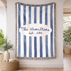 May include: A blue and white striped throw blanket with a fringed edge. The blanket features the text "The Hamiltons est. 2015" in a cursive font within an oval shape. The blanket is hanging on a white wall.