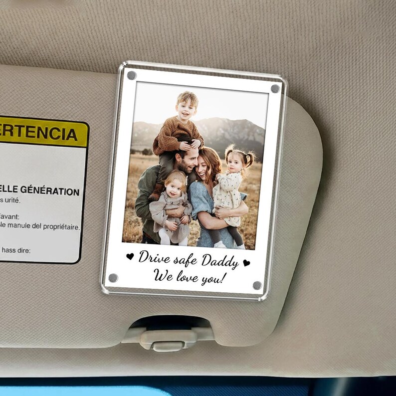 May include: A clear acrylic photo frame with a family photo, attached to a car visor. The photo shows a family of five, with the text "Drive safe Daddy, We love you!" in black script.