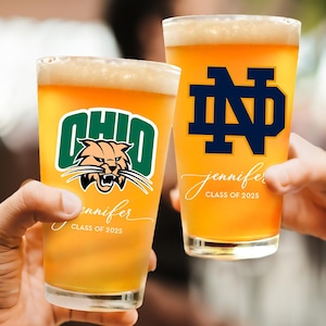 May include: Two pint glasses filled with beer, held by hands. One glass features the Ohio Bobcats logo in green and orange, with the name "Jennifer" and "Class of 2025" in script. The other glass displays the Notre Dame logo in navy blue, with the same name and class year.