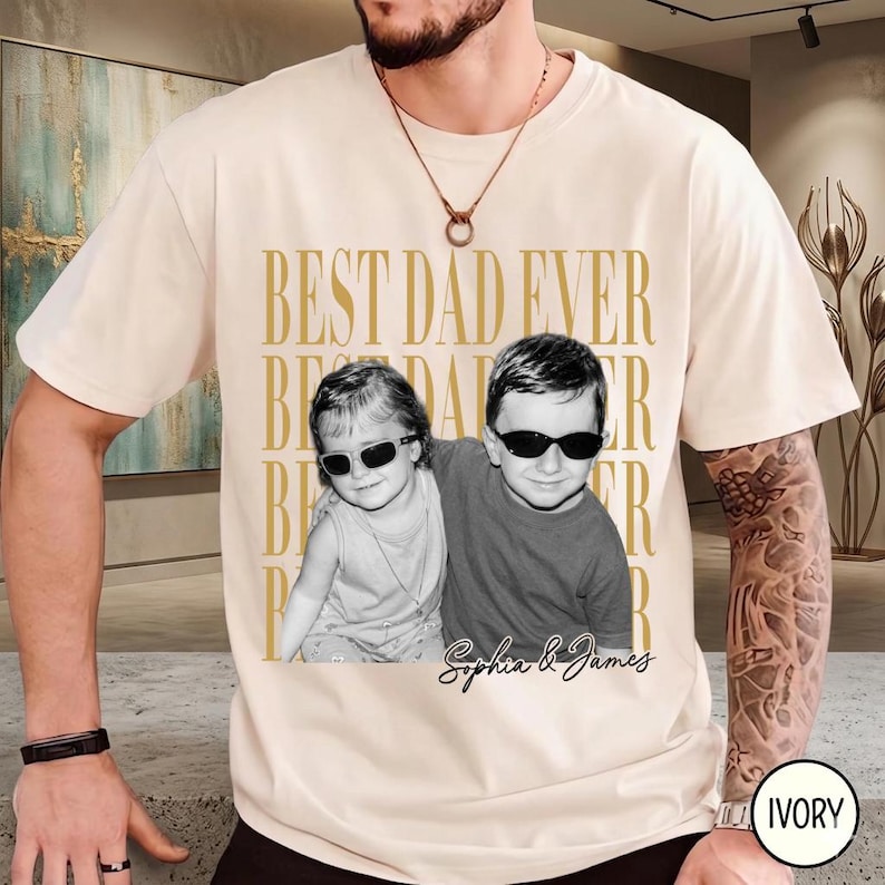 Pode incluir: Camiseta cor marfim com o texto dourado "BEST DAD EVER". Apresenta uma foto em preto e branco de duas crian&ccedil;as usando &oacute;culos de sol, com os nomes "Sophia & James" abaixo. &Eacute; uma pe&ccedil;a de roupa casual.