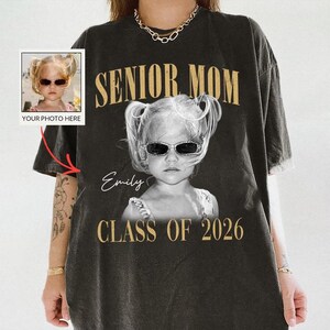 May include: Black t-shirt with the words "SENIOR MOM" in gold lettering. A black and white photo of a child wearing sunglasses is printed on the shirt, with the name "Emily" and "CLASS OF 2026" below.