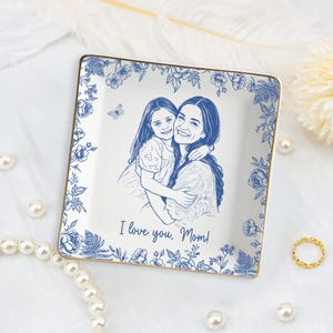 Puede incluir: Plato cuadrado de cer&aacute;mica con borde floral azul y ribete dorado. El centro presenta un boceto azul de una madre y un ni&ntilde;o con el texto "I love you, Mom!". Tambi&eacute;n se incluye una peque&ntilde;a mariposa.