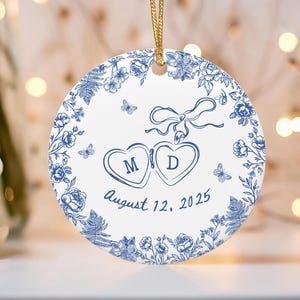 Puede incluir: Adorno redondo de cer&aacute;mica blanca con dise&ntilde;o floral azul y cord&oacute;n dorado. Presenta dos corazones con las iniciales "M" y "D", y la fecha "12 de agosto de 2025".
