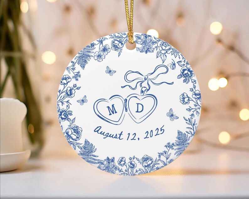 Puede incluir: Adorno redondo de cer&aacute;mica blanca con dise&ntilde;o floral azul y cord&oacute;n dorado. Presenta dos corazones con las iniciales "M" y "D", y la fecha "12 de agosto de 2025".