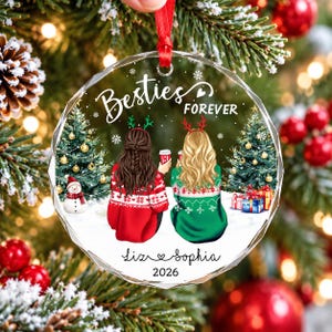 May include: A round, faceted glass Christmas ornament featuring an illustration of two people with long hair wearing holiday sweaters and reindeer antlers. The text reads Besties FOREVER, Liz, Sophia, and 2026, with snowy trees and gifts.