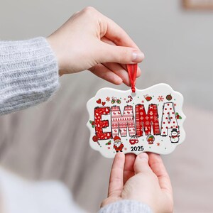 May include: A white ceramic Christmas ornament with a red ribbon. The ornament features the name "EMMA" in a festive red sweater pattern, with holiday icons and the year "2025".
