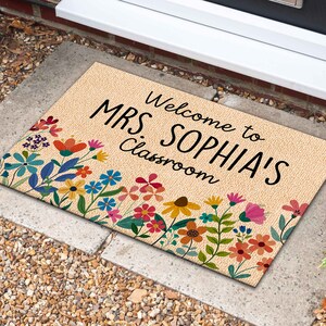 Może przedstawiać: Prostokątna wycieraczka z beżowym tłem i kwiatowym obramowaniem. Mata ma czarny napis "Welcome to MRS. SOPHIA'S Classroom". Kwiatowe obramowanie zawiera kolorowe kwiaty w odcieniach czerwieni, r&oacute;żu, błękitu i ż&oacute;łci.