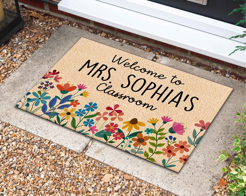 Może przedstawiać: Prostokątna wycieraczka z beżowym tłem i kwiatowym obramowaniem. Mata ma czarny napis "Welcome to MRS. SOPHIA'S Classroom". Kwiatowe obramowanie zawiera kolorowe kwiaty w odcieniach czerwieni, r&oacute;żu, błękitu i ż&oacute;łci.