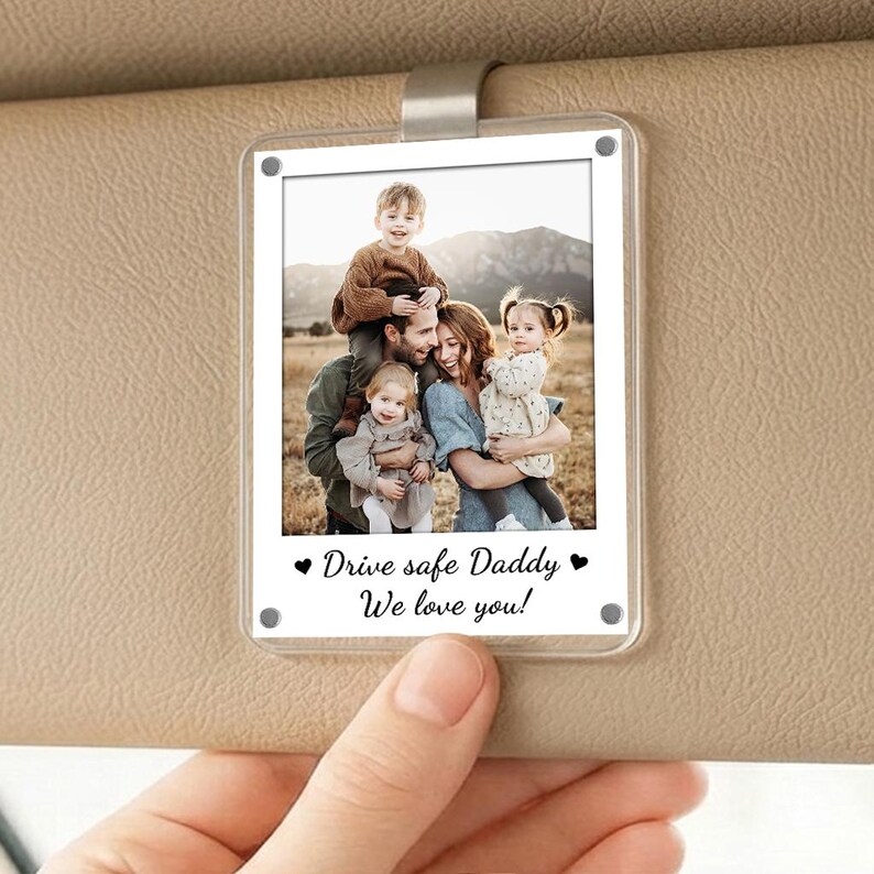 May include: Clear acrylic car visor photo holder with a family photo. The photo features a family of five with the text "Drive safe Daddy We love you!" in black. The holder has a silver clip.
