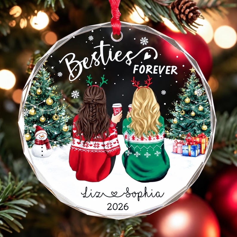May include: A round, faceted glass Christmas ornament featuring an illustration of two people from behind, wearing holiday sweaters and reindeer headbands. The text reads Besties FOREVER, Liz, Sophia, and 2026, with snowy trees and gifts.
