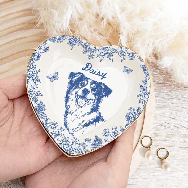 May include: Heart-shaped ceramic trinket dish with a blue and white floral design and a gold rim. The dish features a smiling dog portrait and the name "Daisy" in script. Decorative butterflies are also included.