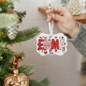 May include: A white ceramic Christmas ornament with the name "EMMA" in red, patterned letters. The ornament features festive designs like sweaters, candy canes, and a snowman. The year "2025" is also displayed. A light blue ribbon is attached for hanging.