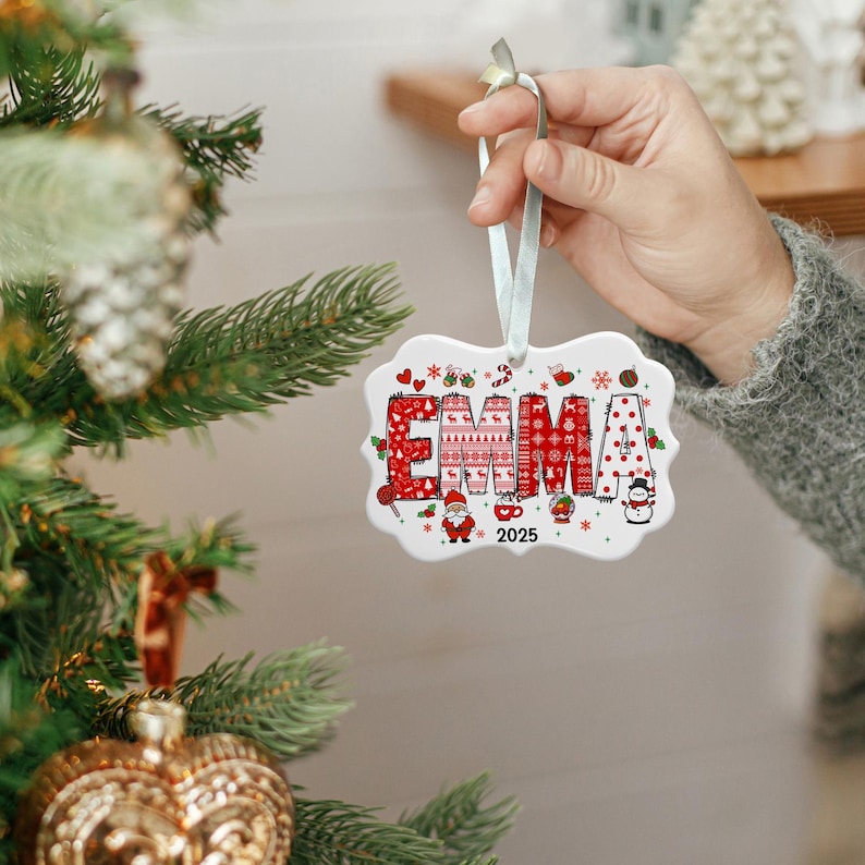 May include: A white ceramic Christmas ornament with the name "EMMA" in red, patterned letters. The ornament features festive designs like sweaters, candy canes, and a snowman. The year "2025" is also displayed. A light blue ribbon is attached for hanging.