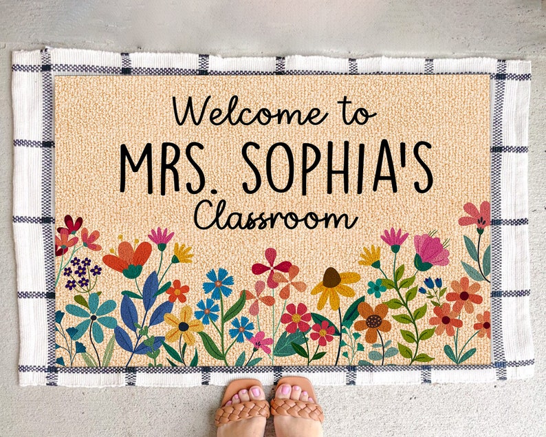 Może przedstawiać: Prostokątna wycieraczka z czarnym napisem "Welcome to MRS. SOPHIA'S Classroom". Mata ma beżowe tło z kwiatową obw&oacute;dką z kolorowymi kwiatami. Obramowanie jest obramowane czarno-białym wzorem w kratę.
