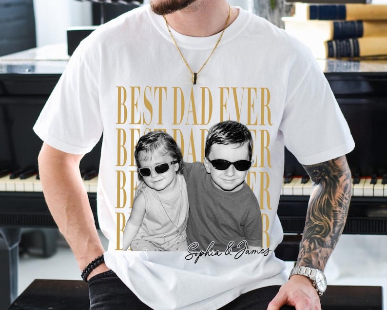 Pode incluir: Camiseta branca com a frase dourada "BEST DAD EVER". A camiseta apresenta uma foto em preto e branco de duas crian&ccedil;as usando &oacute;culos de sol. Os nomes "Sophia & James" est&atilde;o escritos abaixo.