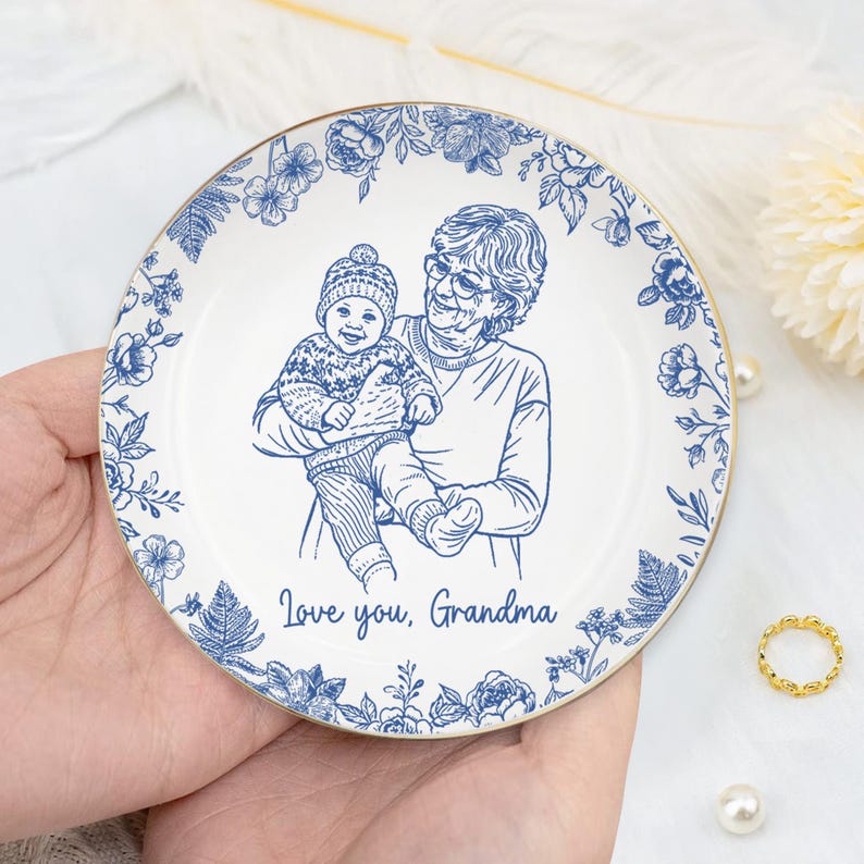 Puede incluir: Un plato de cer&aacute;mica blanco con un borde floral azul y un borde dorado. El centro presenta un dibujo lineal azul de una abuela sosteniendo a un beb&eacute;, con el texto "Love you, Grandma."