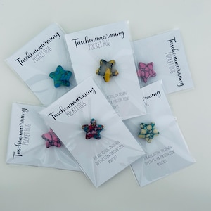 Könnte beinhalten: Eine Auswahl an sternförmigen Taschenumarmungen in verschiedenen Farben, darunter Blau, Gelb und Rosa. Jeder Stern ist auf einer weißen Karte mit dem Text "Taschenumarmung POCKET HUG" verpackt. Die Karten enthalten auch deutschen Text.