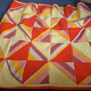 May include: A knitted blanket featuring a geometric pattern of squares composed of triangles. The design uses a colour palette of bright orange, pale yellow, golden yellow, and muted mauve, arranged in a repeating, symmetrical grid layout.