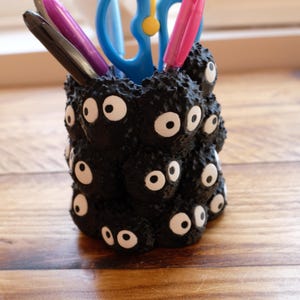 Peut inclure: Un porte-crayon cylindrique noir avec une surface texturée recouverte de plusieurs petites créatures rondes ressemblant à de la suie, dotées de grands yeux blancs et de pupilles noires. Il contient des ciseaux bleus, un stylo rose et un stylo noir.