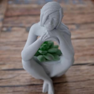 May include: A gray concrete planter shaped like a woman holding a small green succulent plant.