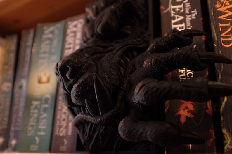 May include: A black wooden bookend shaped like a wolf's head with sharp teeth and claws. The bookend is holding up a stack of books with titles like "Clash of Kings" and "The Wise Man's Fear".