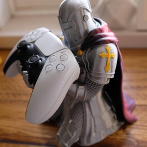 May include: A silver-colored knight figurine in plate armor with a dark red cape and a yellow cross emblem on the shoulder, kneeling while holding a white and black video game controller with circular action buttons and two analog sticks.
