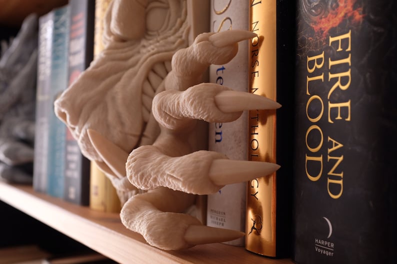 May include: A white dragon claw bookend with sharp, pointed claws holding up a book titled "Norse Mythology" with a gold spine. The bookend is on a bookshelf with other books, including one titled "Fire and Blood" with a black cover and gold lettering.