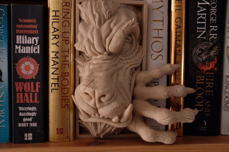 May include: A white ceramic sculpture of a snarling monster with a large mouth and sharp teeth. The sculpture is mounted on a bookshelf between several books, including "Wolf Hall" by Hilary Mantel and "Fire and Blood" by George R.R. Martin.