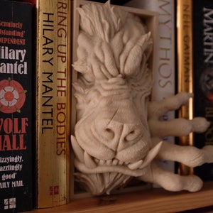 May include: A white ceramic bookend shaped like a monster's head with sharp teeth and claws. The bookend is placed between books on a bookshelf.
