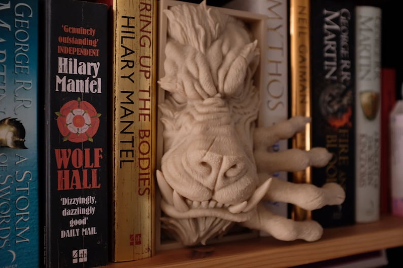 May include: A white ceramic bookend shaped like a monster's head with sharp teeth and claws. The bookend is placed between books on a bookshelf.