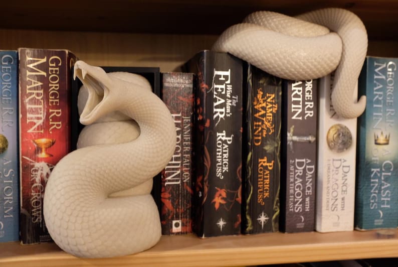 Puede incluir: Un sujetalibros de serpiente blanco de cer&aacute;mica con la boca abierta, sosteniendo una pila de novelas de fantas&iacute;a. Los libros incluyen "A Storm of Swords", "A Feast for Crows", "The Wise Man's Fear", "The Name of the Wind", "A Dance with Dragons" y "Clash of Kings".